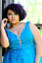 A woman with short, curly purple hair wearing a bright blue lace and tulle formal dress, posing outdoors against a rustic wooden post with a blurred green background.