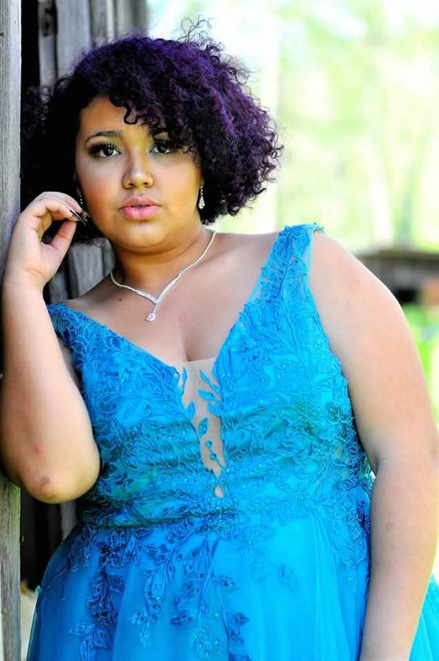 A woman with short, curly purple hair wearing a bright blue lace and tulle formal dress, posing outdoors against a rustic wooden post with a blurred green background.