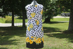 A sleeveless white dress with black polka dots and yellow floral prints, displayed on a mannequin outdoors in a grassy park area with trees in the background.