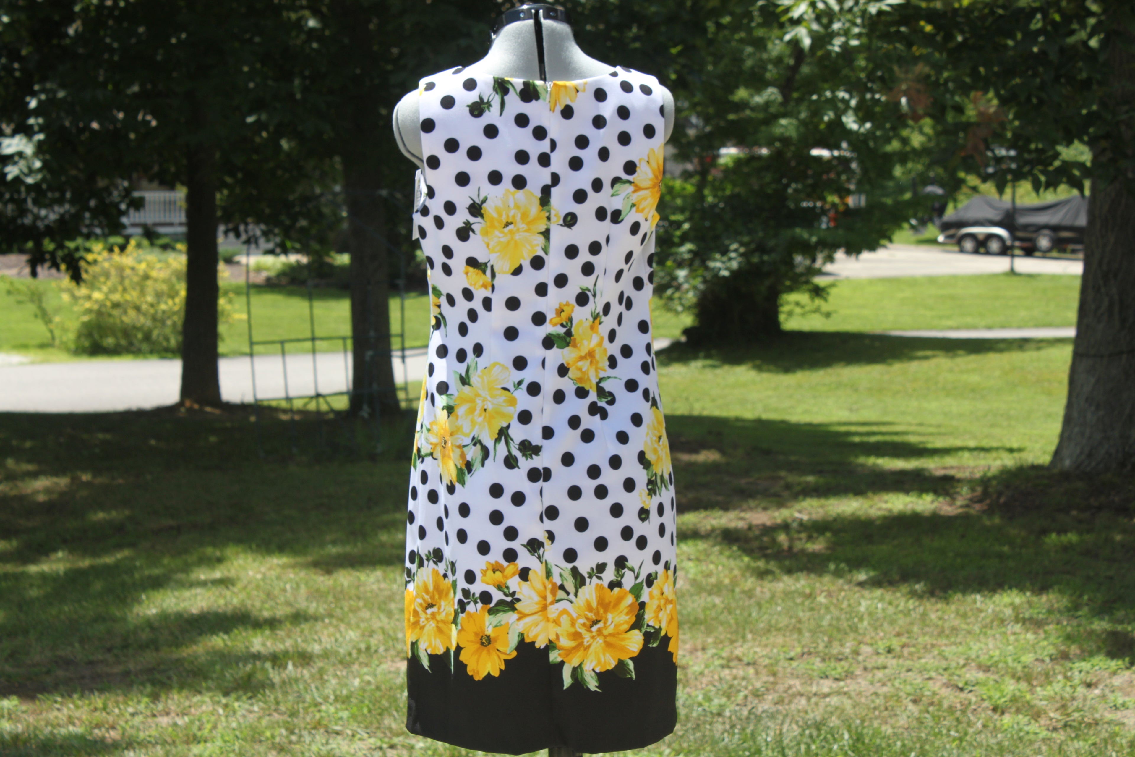A sleeveless white dress with black polka dots and yellow floral prints, displayed on a mannequin outdoors in a grassy park area with trees in the background.