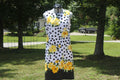 A sleeveless white dress with black polka dots and yellow floral prints, displayed on a mannequin outdoors in a grassy yard with trees.