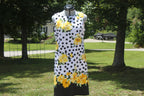 A sleeveless white dress with black polka dots and yellow floral prints, displayed on a mannequin outdoors in a grassy yard with trees.