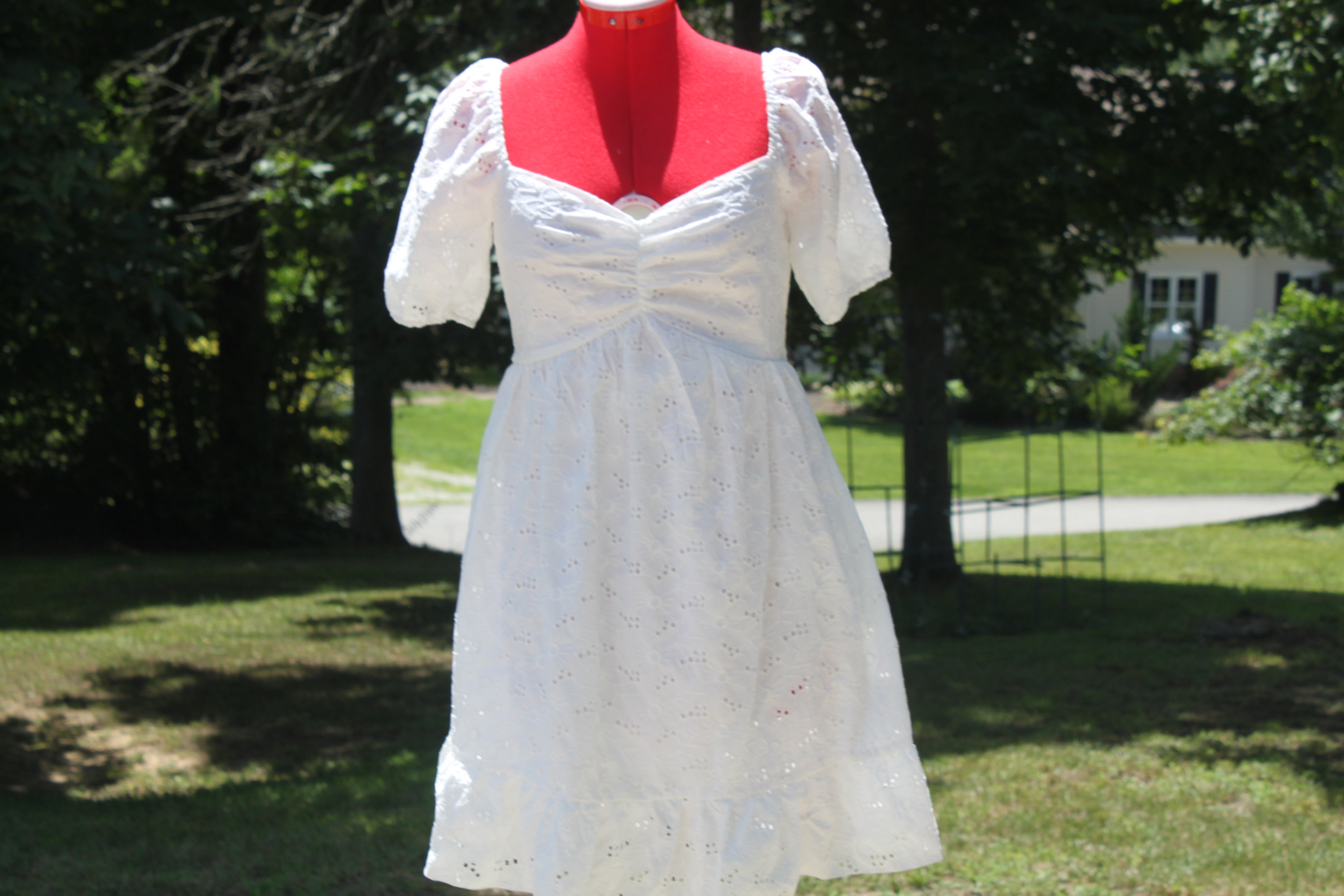 A white eyelet mini dress with puff sleeves and a sweetheart neckline displayed on a red mannequin outdoors in a grassy yard with trees.