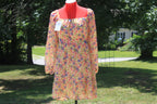 A long-sleeve floral midi dress displayed on a red mannequin outdoors in a grassy yard with trees and a house in the background. The dress has a warm peach base with a multicolor floral print in pink, purple, and blue, and a square neckline.