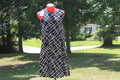 A sleeveless black dress with a white geometric lattice pattern displayed on a red mannequin outdoors in a grassy yard with trees.