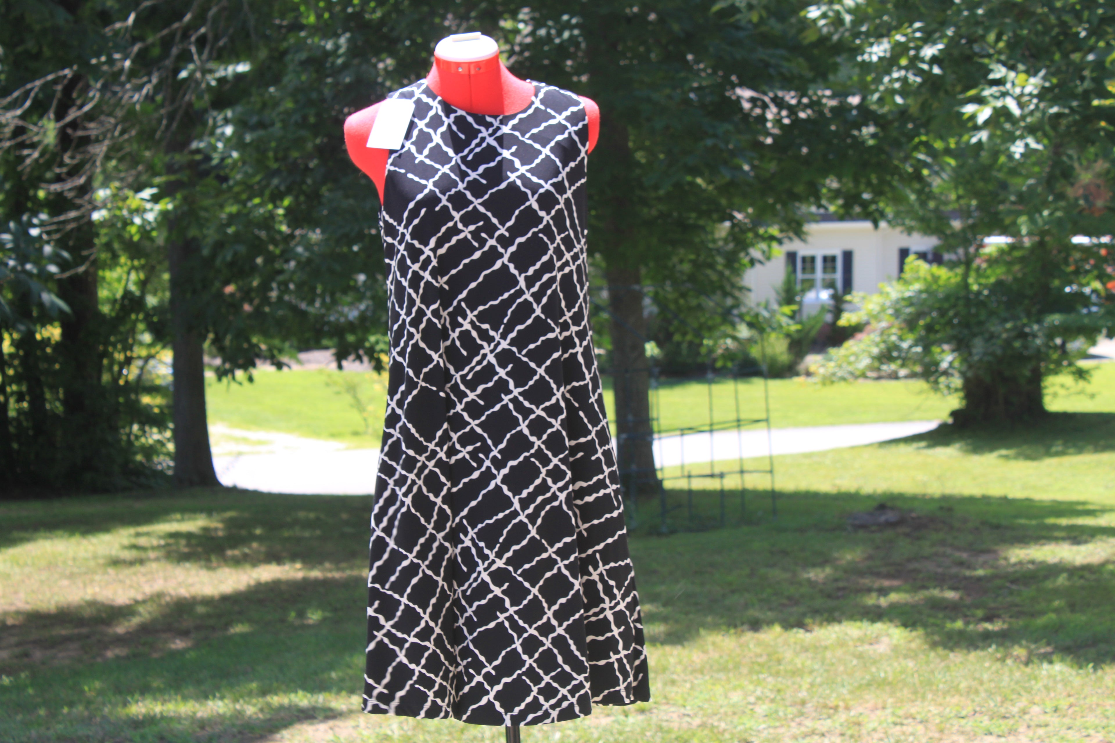 A sleeveless black dress with a white geometric lattice pattern displayed on a red mannequin outdoors in a grassy yard with trees.