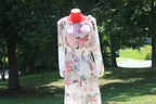 A long-sleeve white maxi dress with a pink and green floral print, displayed on a red mannequin outdoors in a grassy park area with trees.
