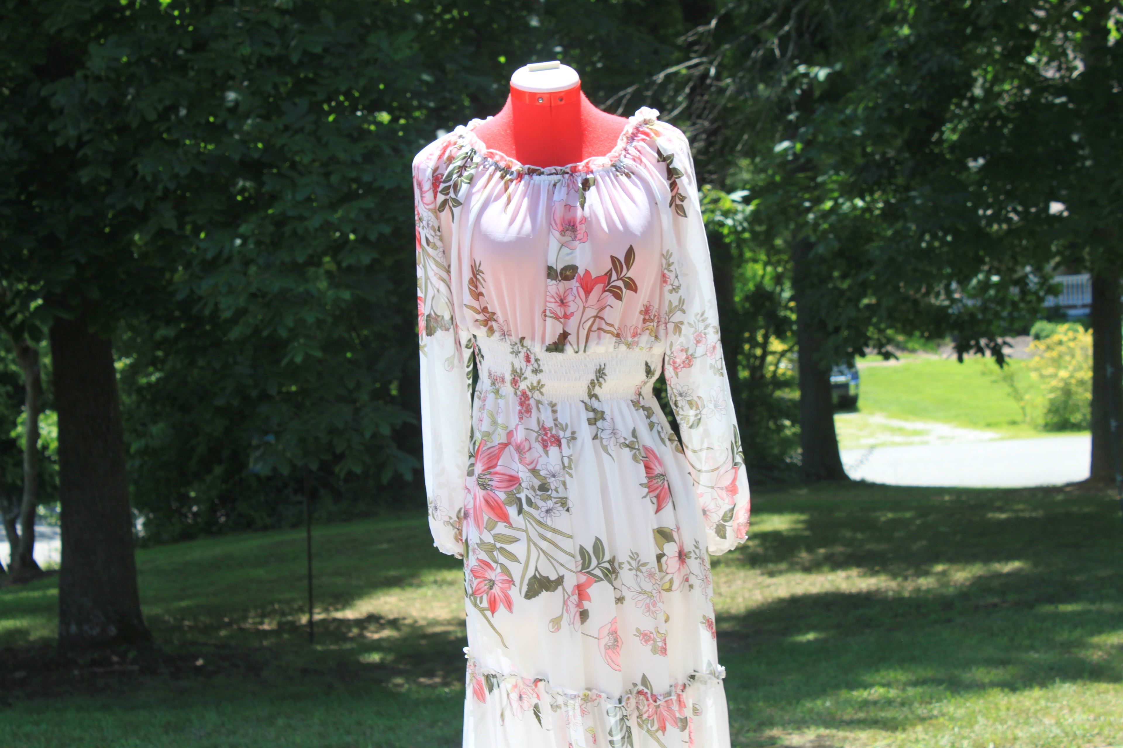 A long-sleeve white maxi dress with a pink and green floral print, displayed on a red mannequin outdoors in a grassy park area with trees.