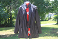 A dark Black hooded jacket displayed on a mannequin outdoors, layered over a red quilted vest and a red top. The background shows a blurred green lawn and tree