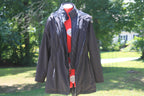 A dark Black hooded jacket displayed on a mannequin outdoors, layered over a red quilted vest and a red top. The background shows a blurred green lawn and tree