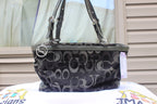 A Coach handbag with a black and gray monogram pattern, featuring dual shoulder straps, a top zipper, and a silver-tone logo charm, displayed on a white surface with a window and siding in the background.