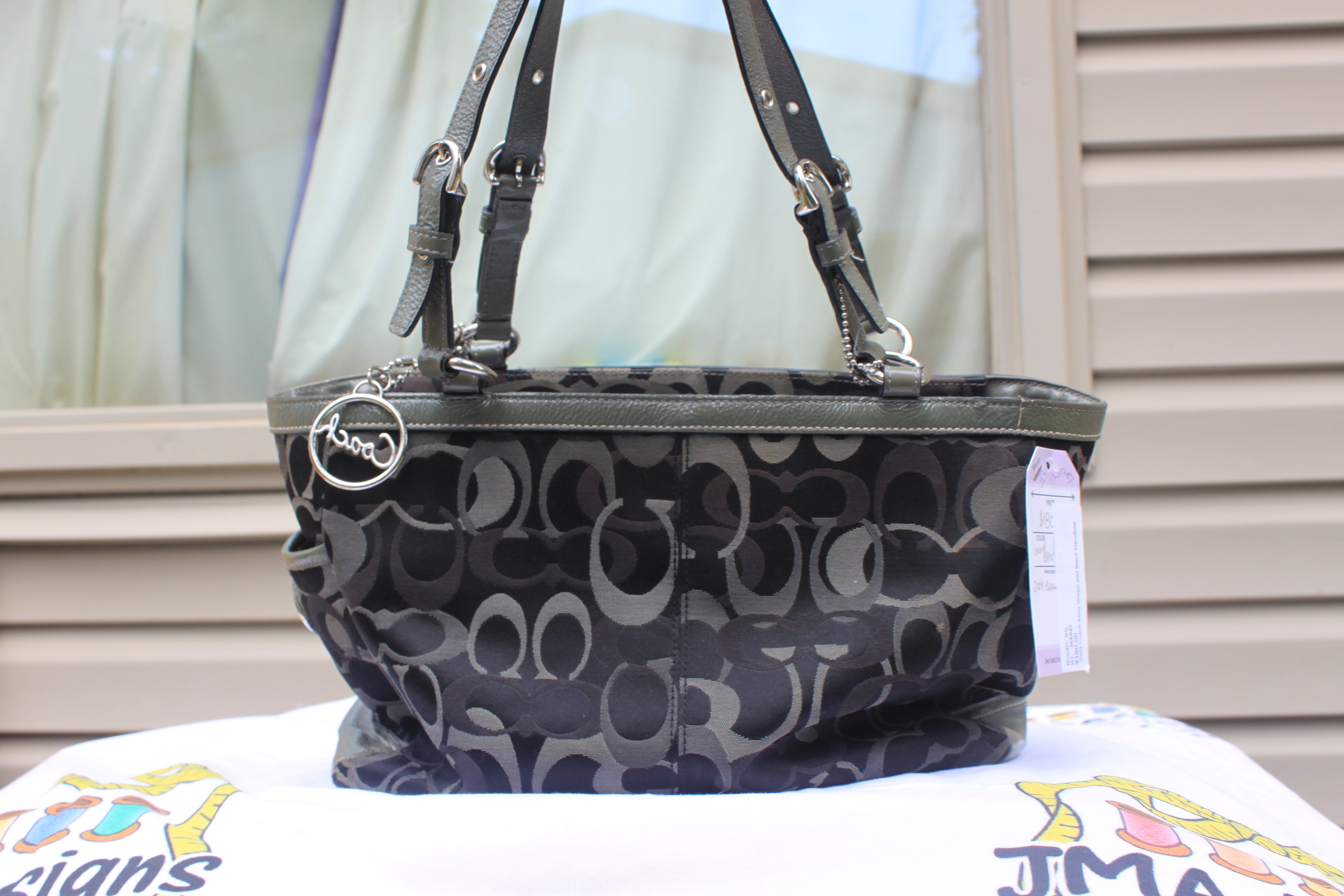 A Coach handbag with a black and gray monogram pattern, featuring dual shoulder straps, a top zipper, and a silver-tone logo charm, displayed on a white surface with a window and siding in the background.