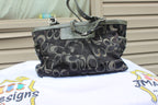 A Coach canvas shoulder bag with a signature monogram pattern in black, gray, and beige, featuring dark olive green leather trim and handles, displayed on a white printed cloth.
