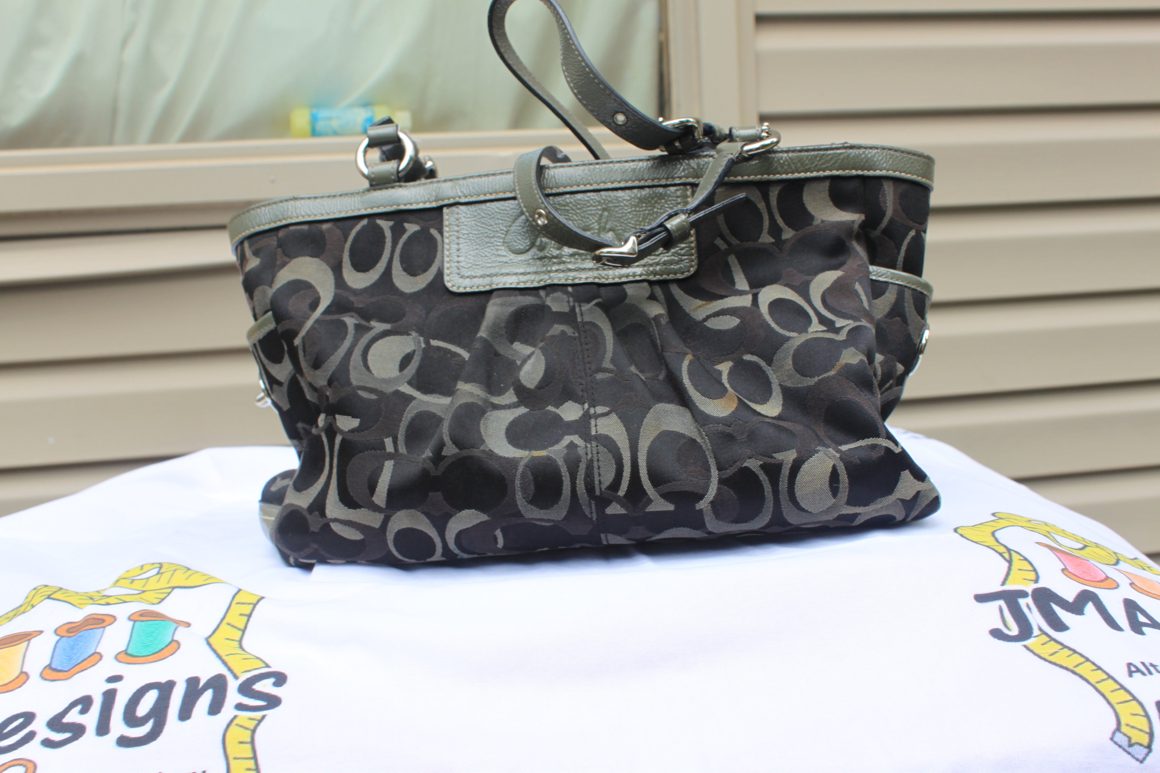 A Coach canvas shoulder bag with a signature monogram pattern in black, gray, and beige, featuring dark olive green leather trim and handles, displayed on a white printed cloth.