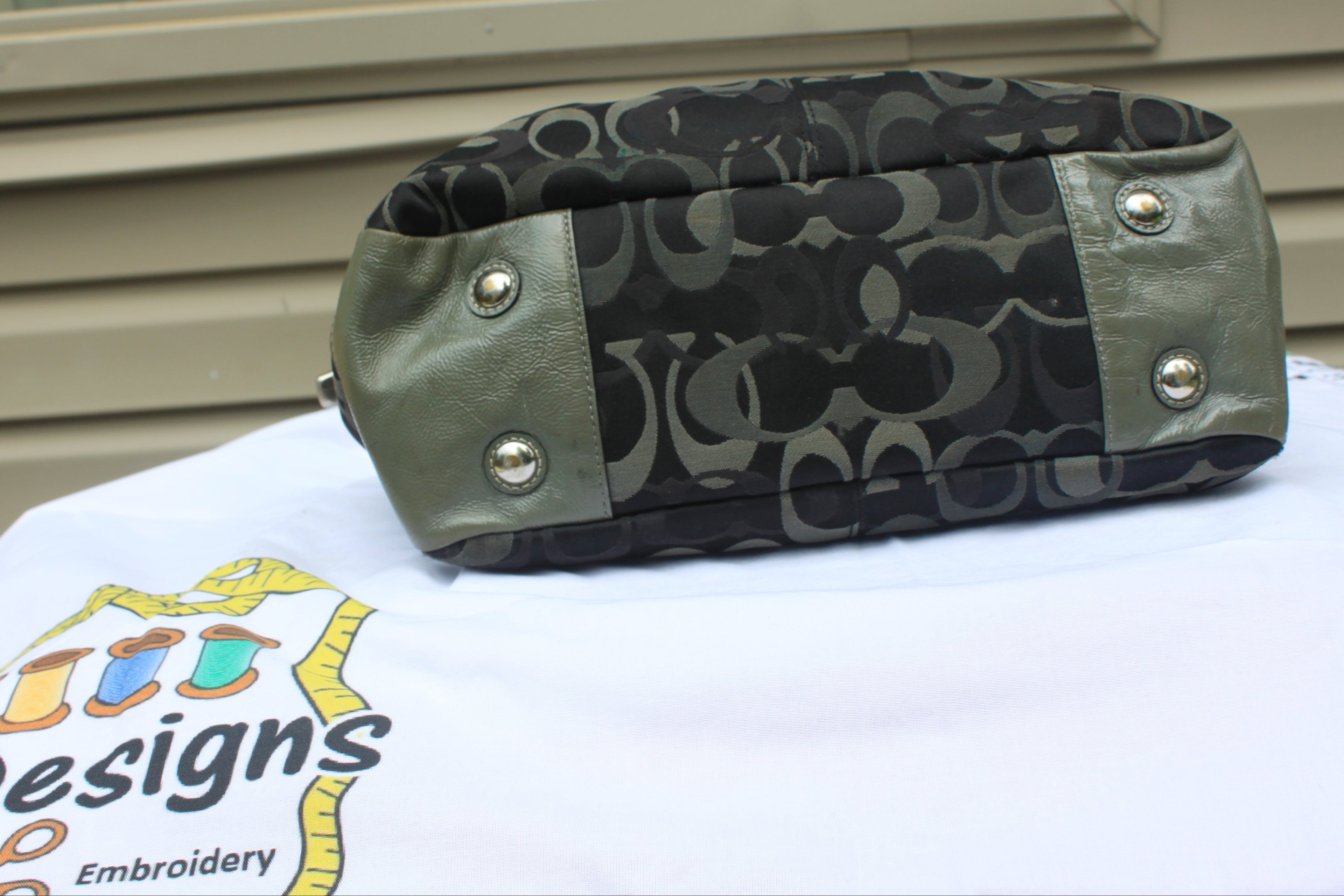 A Coach canvas and leather shoulder bag with the signature 'C' monogram pattern, olive green leather trim, and silver-tone snap buttons, resting on a white embroidered T-shirt.