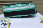 A cylindrical duffel bag with horizontal stripes of green, white, and black, resting on a white embroidered tablecloth in front of a beige siding wall.