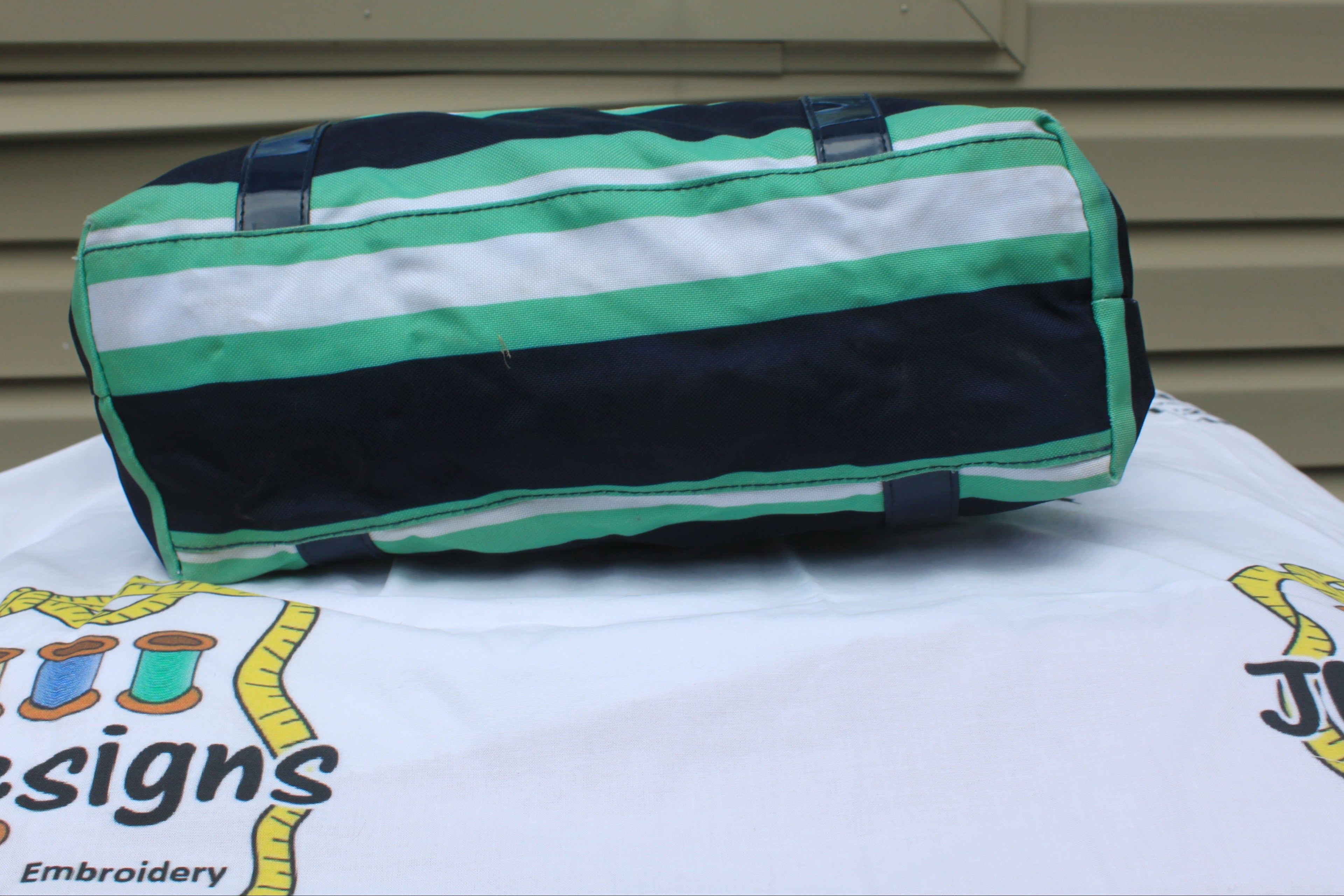 A cylindrical duffel bag with horizontal stripes of green, white, and black, resting on a white embroidered tablecloth in front of a beige siding wall.