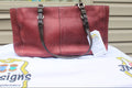 A burgundy pebbled leather handbag with dark brown shoulder straps and silver-tone hardware, resting on a white embroidered tablecloth with a product tag attached.