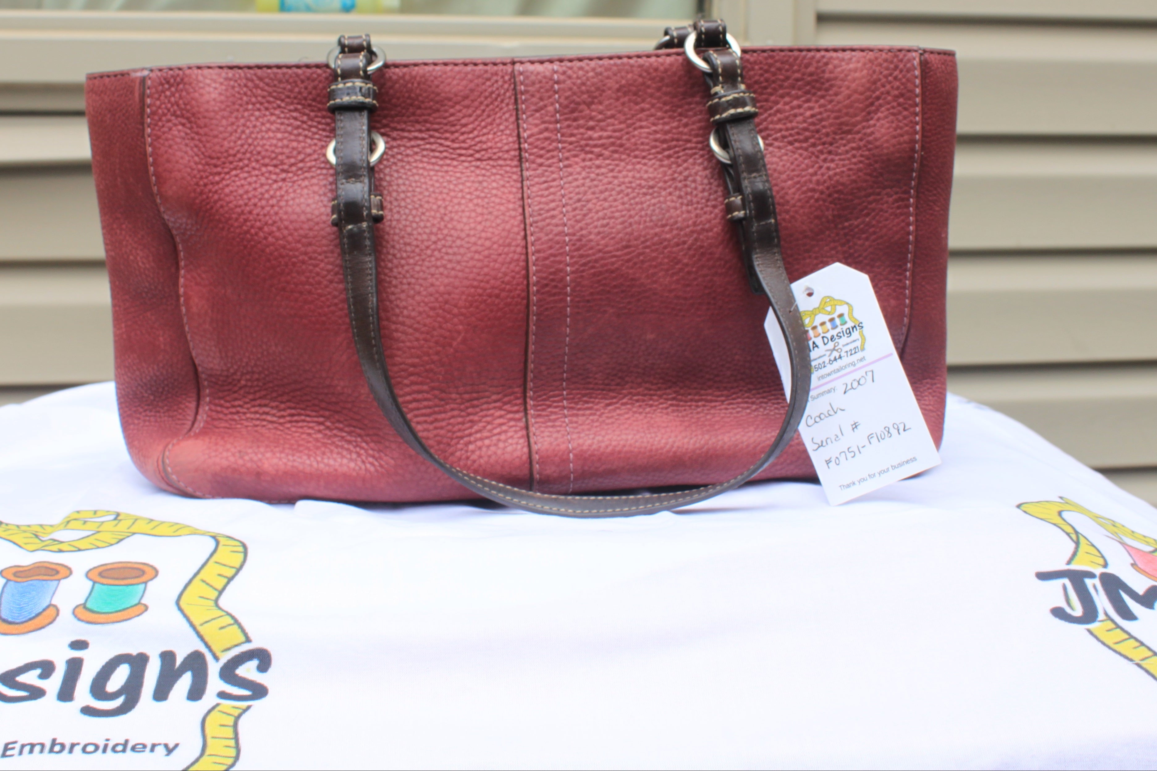 A burgundy pebbled leather handbag with dark brown shoulder straps and silver-tone hardware, resting on a white embroidered tablecloth with a product tag attached.