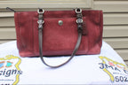 A burgundy pebbled leather handbag with dark brown leather straps and silver-tone hardware, displayed on a white embroidered tablecloth in front of a beige siding wall.