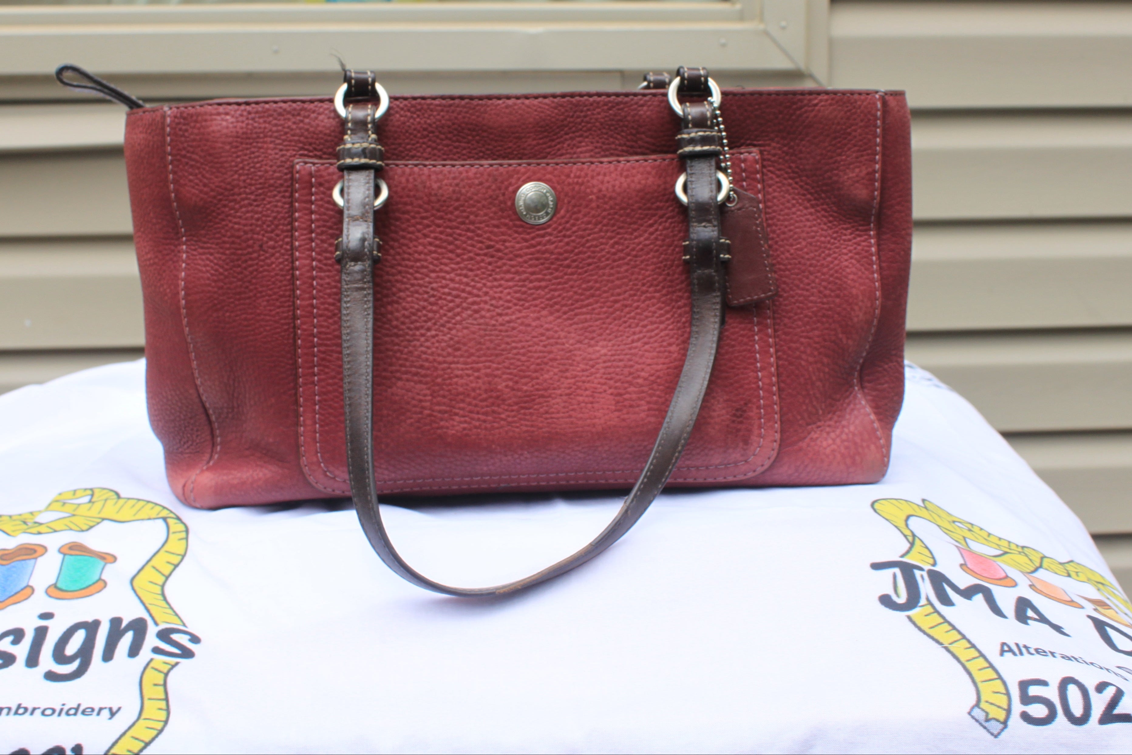 A burgundy pebbled leather handbag with dark brown leather straps and silver-tone hardware, displayed on a white embroidered tablecloth in front of a beige siding wall.