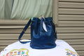 A blue leather bucket bag with a drawstring top and shoulder strap, displayed on a white printed blanket in front of a beige siding wall.