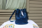 A blue leather bucket bag with a drawstring top and shoulder strap, displayed on a white printed blanket in front of a beige siding wall.