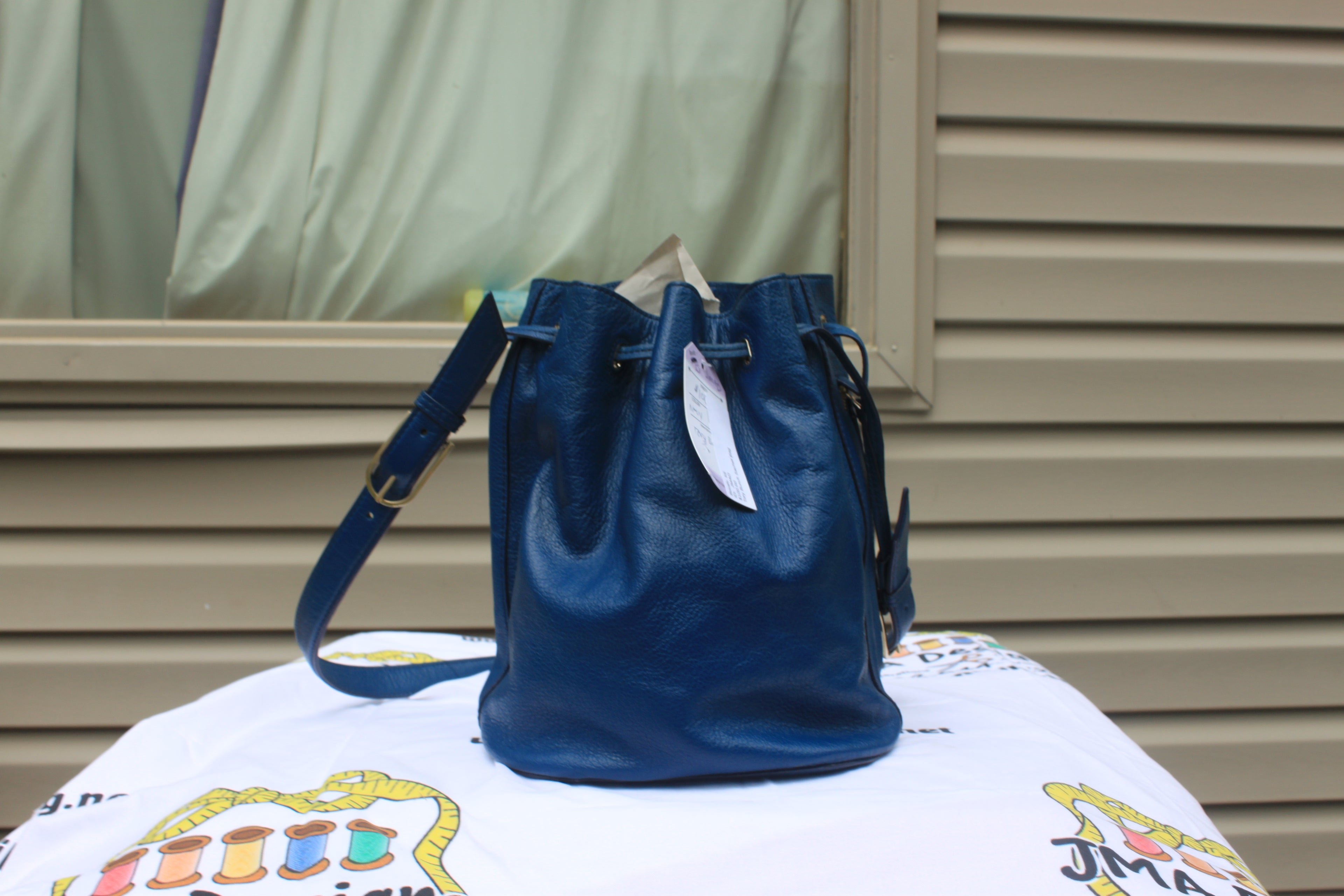 A blue leather bucket bag with a drawstring top and shoulder strap, displayed on a white printed blanket in front of a beige siding wall.