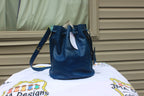 A dark blue leather bucket bag with a drawstring top and a shoulder strap, resting on a white printed blanket in front of a beige house siding background.