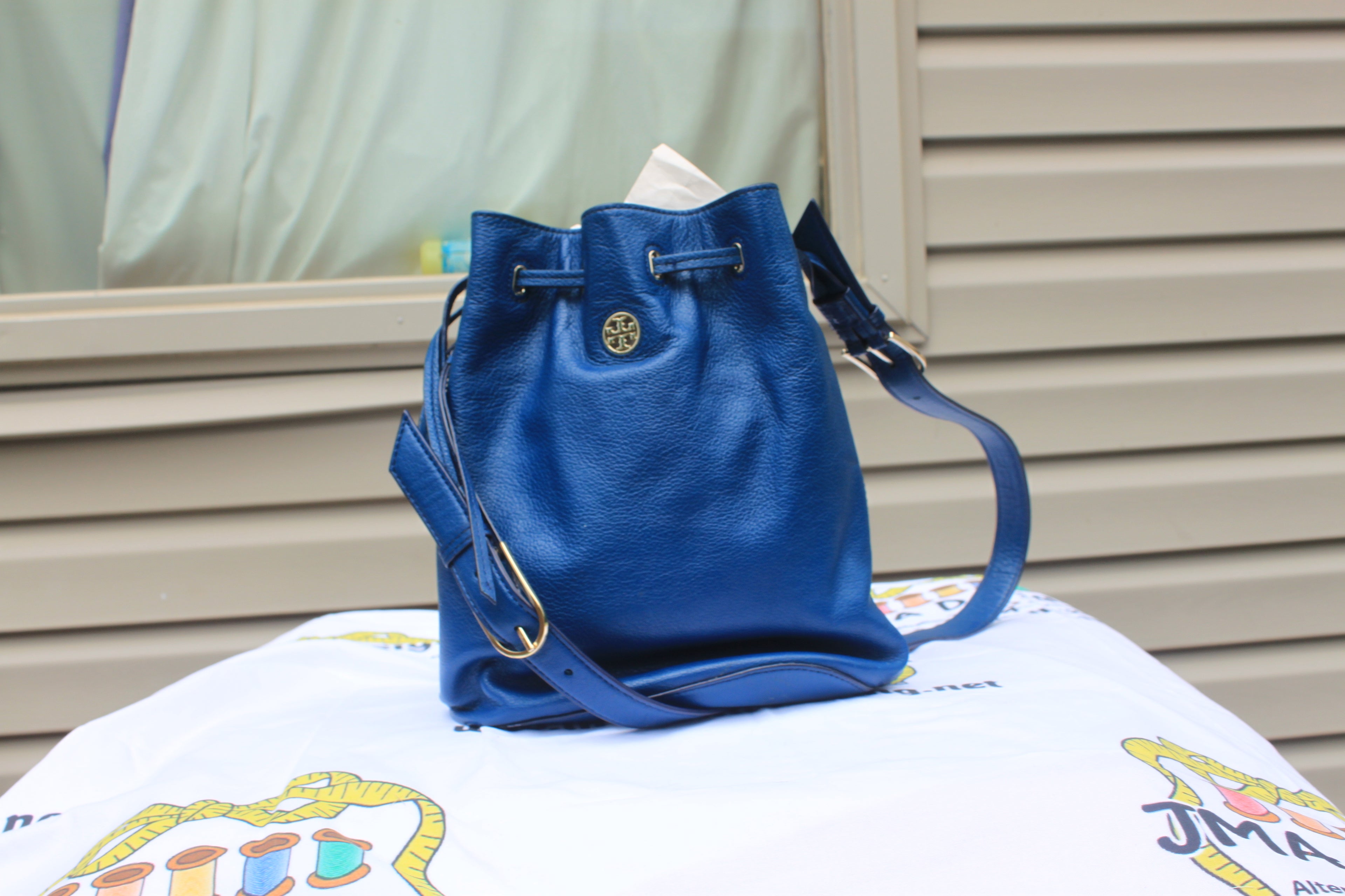 A blue pebbled leather bucket bag with a drawstring closure and a gold-tone circular logo medallion, displayed on a white printed blanket in front of a beige slatted wall.