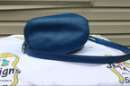 A blue leather crossbody bag with a matching strap, resting on a white embroidered blanket in front of a beige house siding background.