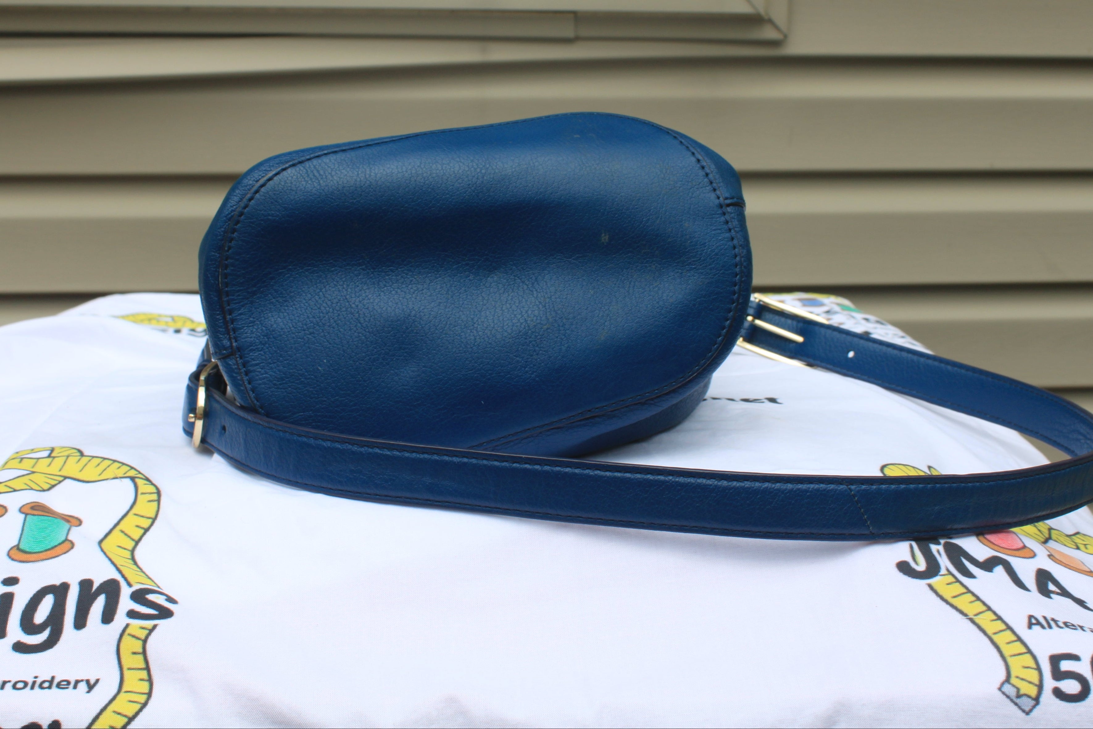 A blue leather crossbody bag with a matching strap, resting on a white embroidered blanket in front of a beige house siding background.