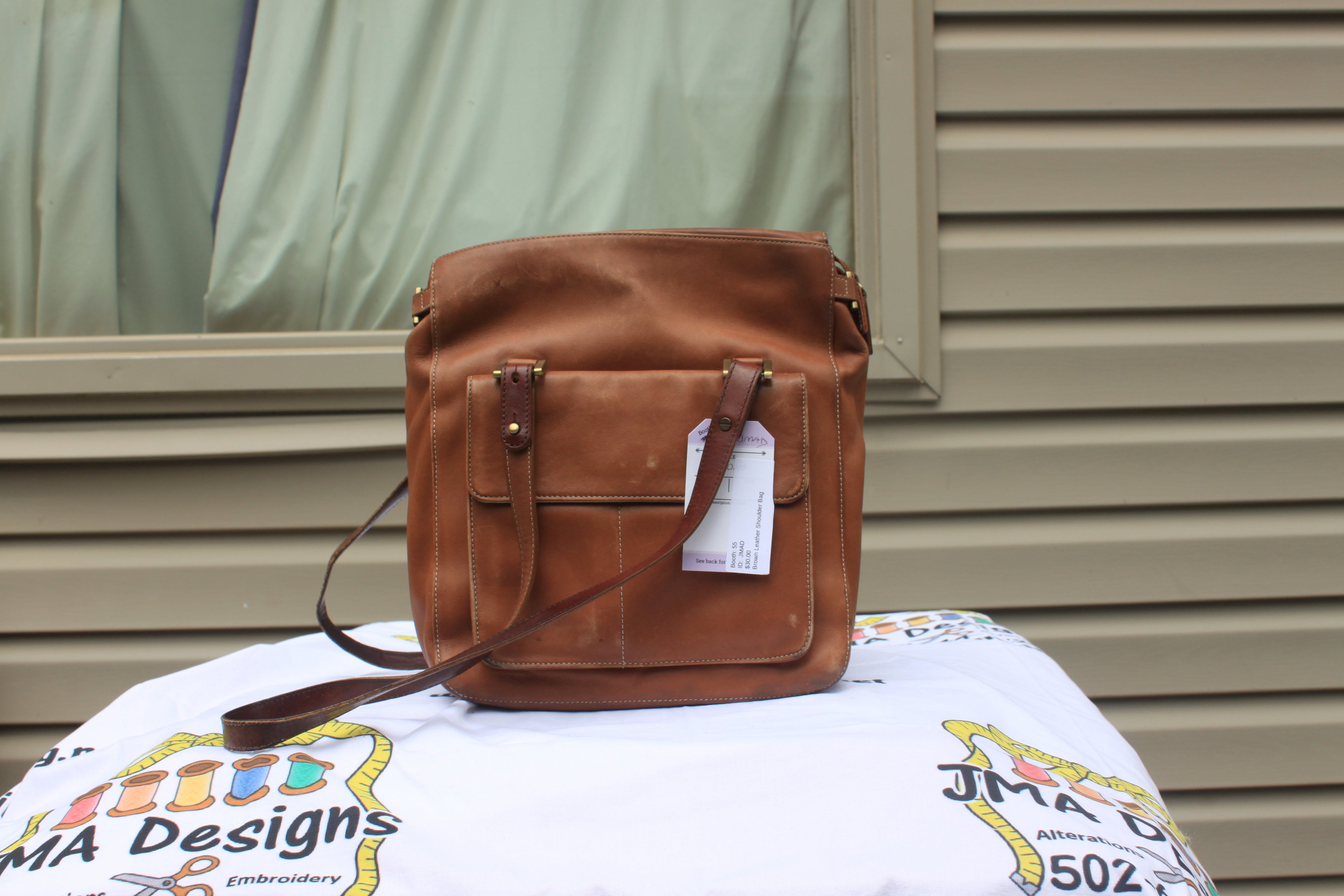 A brown leather crossbody bag with a front pocket and a white product tag, displayed on a white printed blanket in front of a beige house exterior.