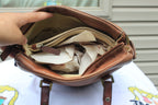 A brown leather shoulder bag shown open, with a hand pointing inside at multiple cream-colored zippered pouches and paper filler. The bag is resting on a white printed surface with a house siding background.