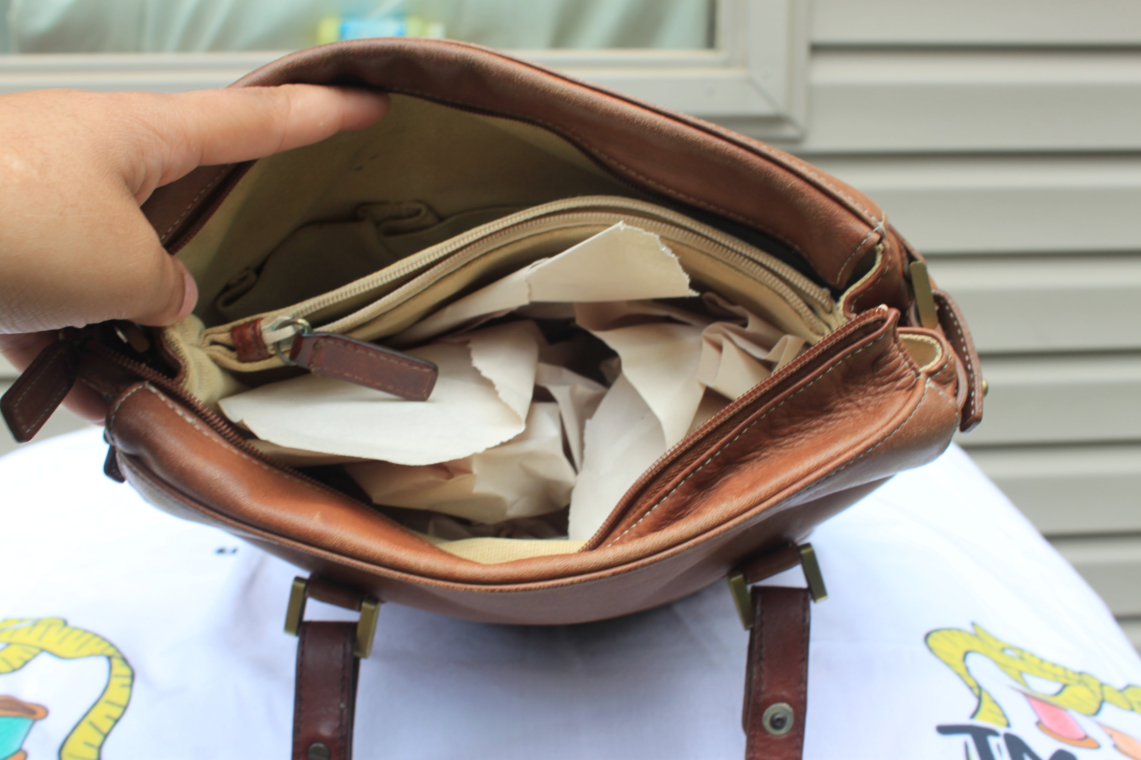 A brown leather shoulder bag shown open, with a hand pointing inside at multiple cream-colored zippered pouches and paper filler. The bag is resting on a white printed surface with a house siding background.