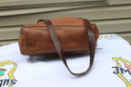 A brown leather shoulder bag with a matching strap, resting on a white printed blanket in front of a beige house siding wall.