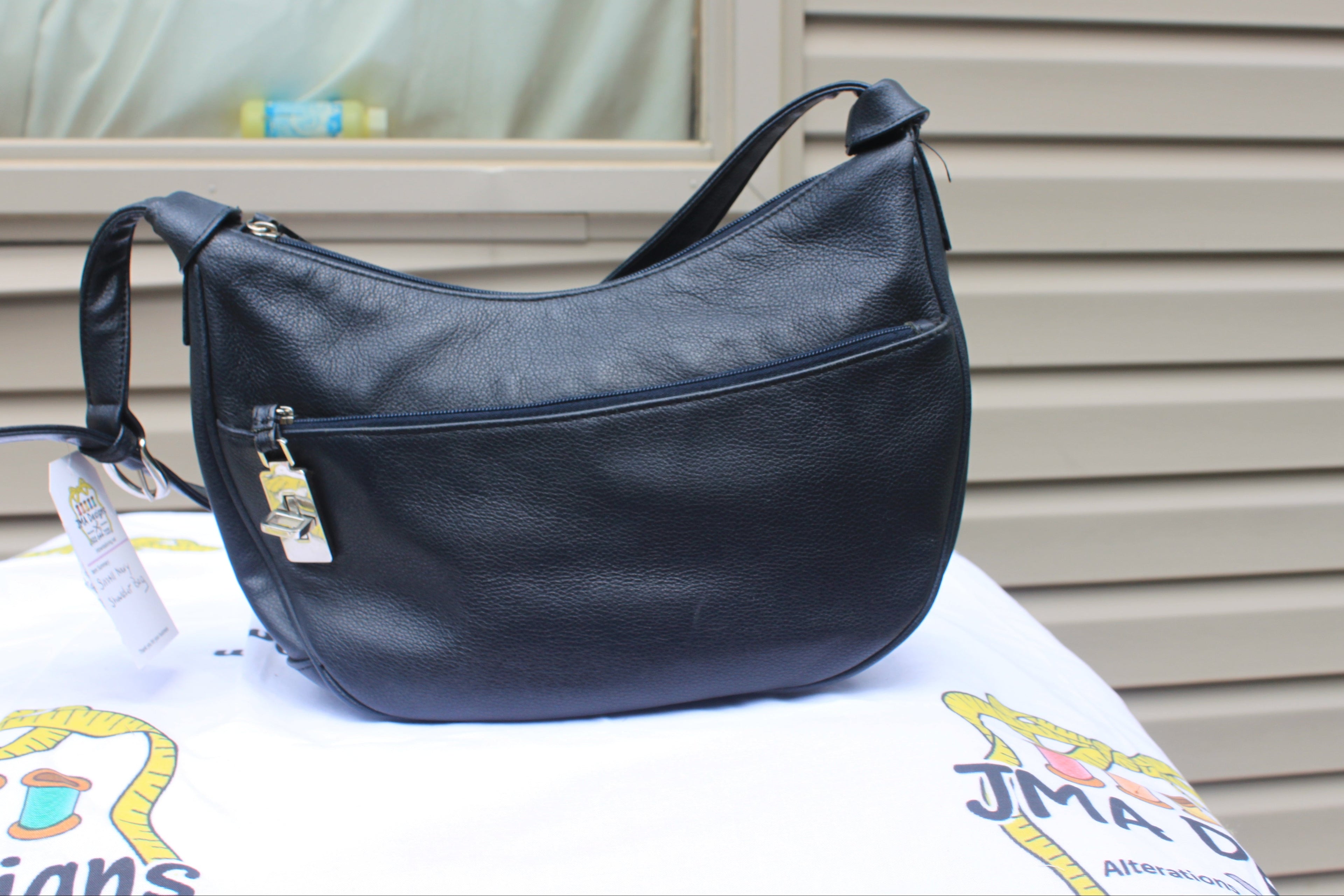 A black faux-leather crescent-shaped shoulder bag with a front zippered pocket and a hanging product tag, displayed on a white surface in front of a beige siding background.