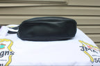 A black faux-leather crossbody bag with a zippered closure, resting on a white printed cloth in front of a beige house siding wall.