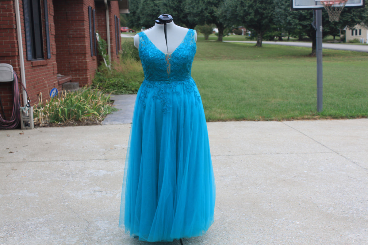 A bright turquoise blue formal gown displayed on a mannequin outdoors on a concrete driveway, with a brick house, grass, trees, and a basketball hoop in the background.