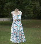 A sleeveless white midi dress with a colorful floral print, displayed on a mannequin outdoors on a grassy lawn with trees in the background.