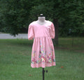 A pink short-sleeve dress with a V-neck and colorful floral print near the hem, displayed on a mannequin outdoors on a grassy lawn with trees in the background.