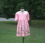 A pink short-sleeve dress with a V-neck and colorful floral print near the hem, displayed on a mannequin outdoors on a grassy lawn with trees in the background.