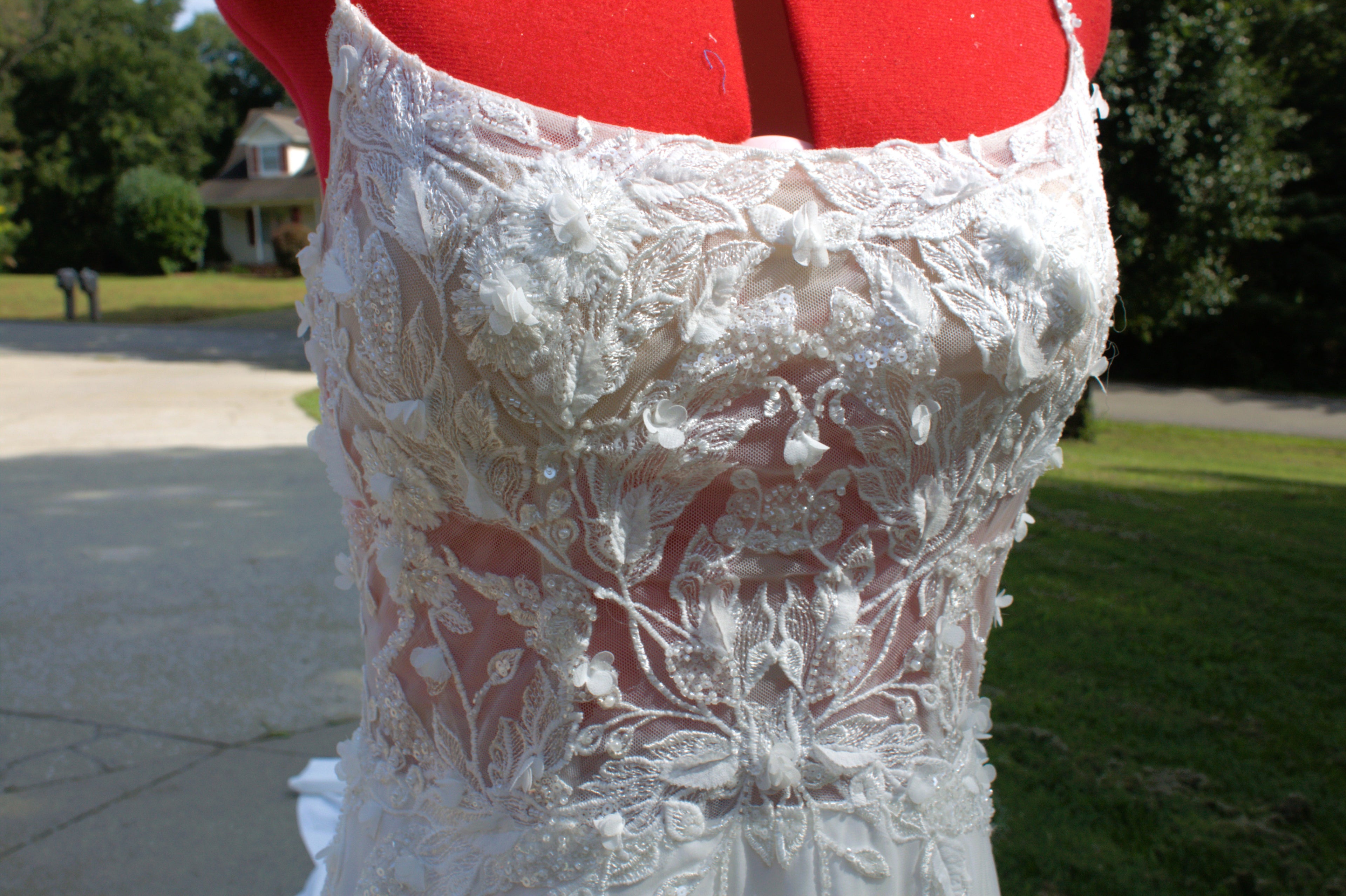 White lace wedding dress bodice displayed on a red dress form outdoors, with a blurred lawn and house in the background.